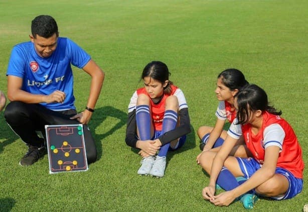 A coach kneeling with a tactics board, talking to three players sitting on the grass