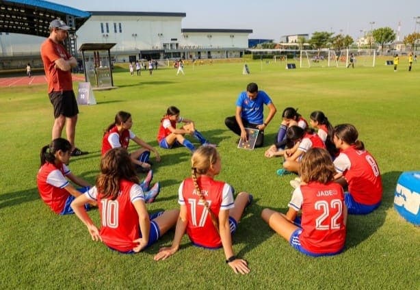 The squad sitting on the grass in a circle listening to their coaches